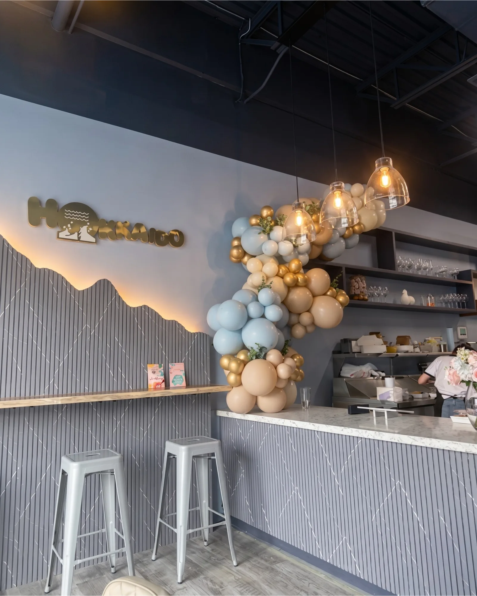 Hokkaido Cake shop interior with gold logo on mountain-slat wall, cream and dusty blue balloon arch, pendant lights, and marble counter