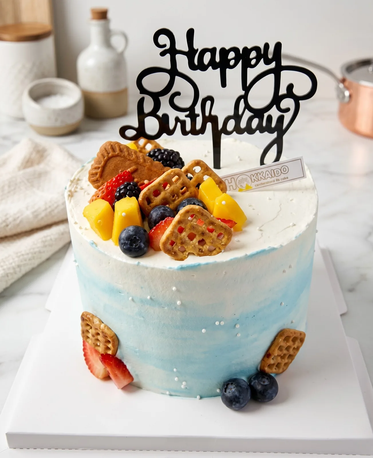 Blue-ombre Happy Birthday cake topped with fresh strawberries, mango, blueberries and biscuits